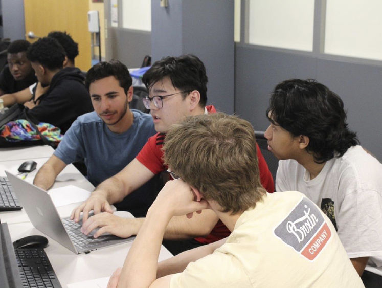 Four students huddle around a laptop in a computer lab