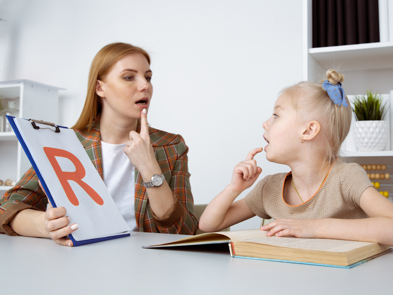 A speech therapist assisting a young girl with her pronunciation, the therapist holding a flashcard with the letter 'R', both pointing at their mouths, book, classroom setting