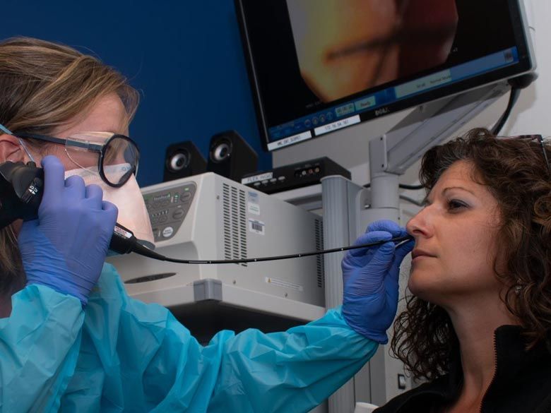 A speech pathologist in full protective gear, including a mask and gloves, administering a nasal endoscopy on a female patient. The patient's face is turned to the side as the endoscope is gently inserted into her nostril. A monitor in the background displays the endoscopic view.