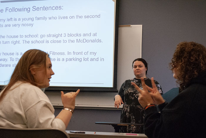 Two students face each other and use sign language, while an instructor watches