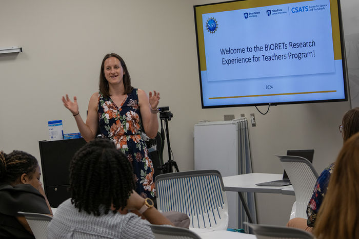 A woman stands in a classroom next to a screen that says "Welcome to the Biorets Research Experience for Teachers Program"