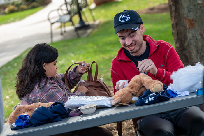 Two students sit at a table outdoors, the materials to stuff a plush animal in front of them