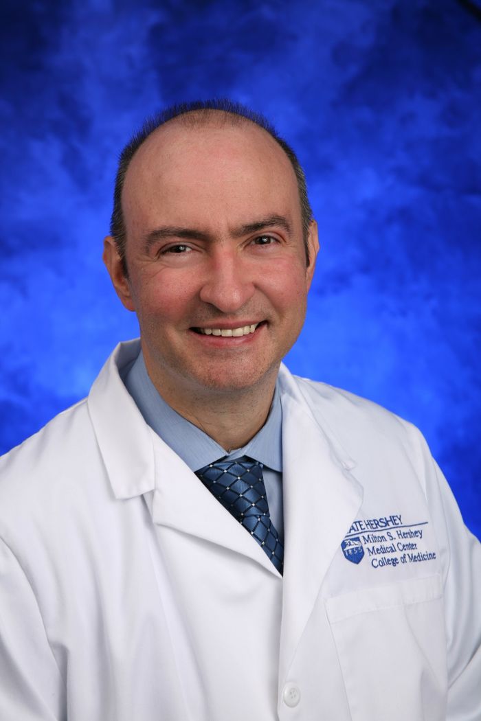 Dr. Sinisa Dovat smiles. He wears a white jacket with the Penn State Health Milton S. Hershey Medical Center and Penn State College of Medicine logo on the breast, a sport shirt and tie.