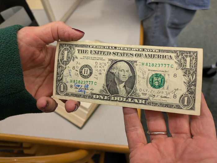 Closeup of hands holding a dollar bill. Dates are written in pen on the dollar bill