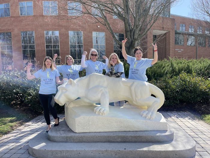 A group of women in matching t-shirts strike muscle poses, standing behind the Lion statue at Penn State Harrisburg