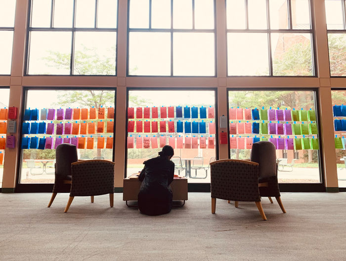 A student sits on the floor at a table in front of colorful bags taped to a large window