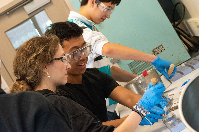 Students wearing safety goggles and blue gloves work with concrete during a civil engineering workshop