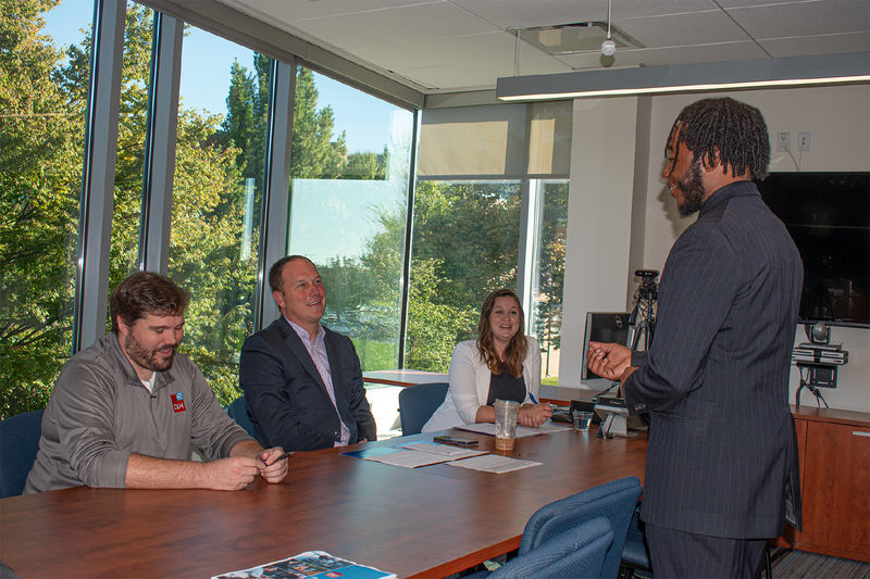 A student in a suit speaks before three company representatives sitting at a table