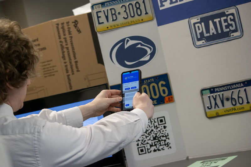 A student holds a phone at arm's length in front of a poster featuring license plates