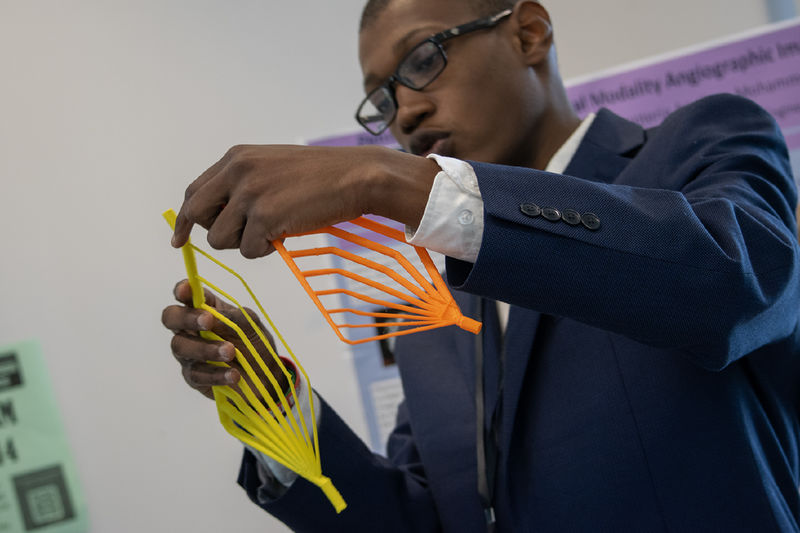 A student in a suit holds two brightly colored objects as he discusses his Capstone project