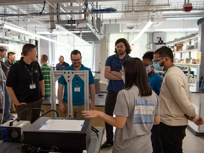 An undergraduate student gives a demo to students of the additive manufacturing and design master's program in an engineering lab with equipment on a table.