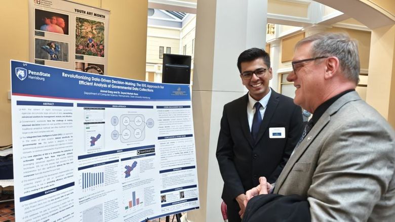 Anmol Garg stands next to his research poster in the state Capitol, while another person looks at the poster