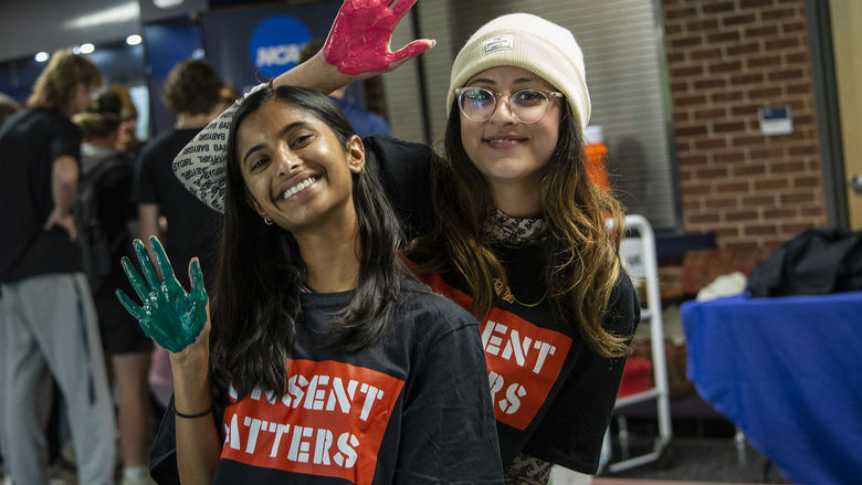 Two students wearing "Consent Matters" shirts hold up their hands, which are painted so they can add their handprints to a banner.
