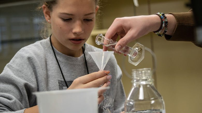 A high school student holds a funnel while another student pours liquid into a container.