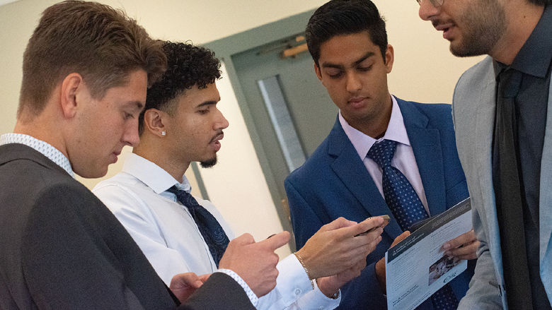 Four students stand together, looking at their phones and holding company brochures