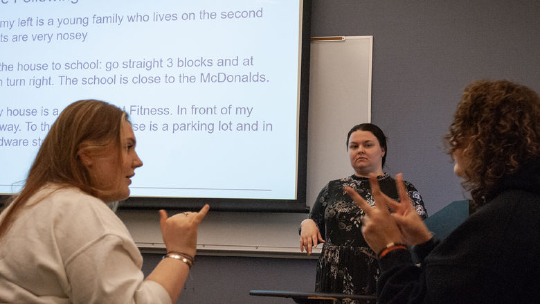 Two students face each other and use sign language, while an instructor watches
