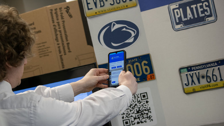 A student holds a phone at arm's length in front of a poster featuring license plates