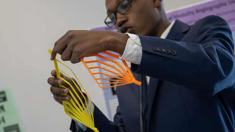 A student in a suit holds two brightly colored objects as he discusses his Capstone project