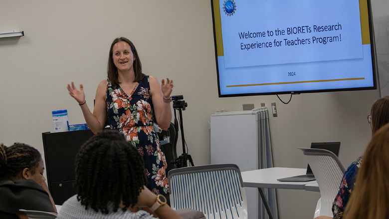 A woman stands in a classroom next to a screen that says "Welcome to the Biorets Research Experience for Teachers Program"