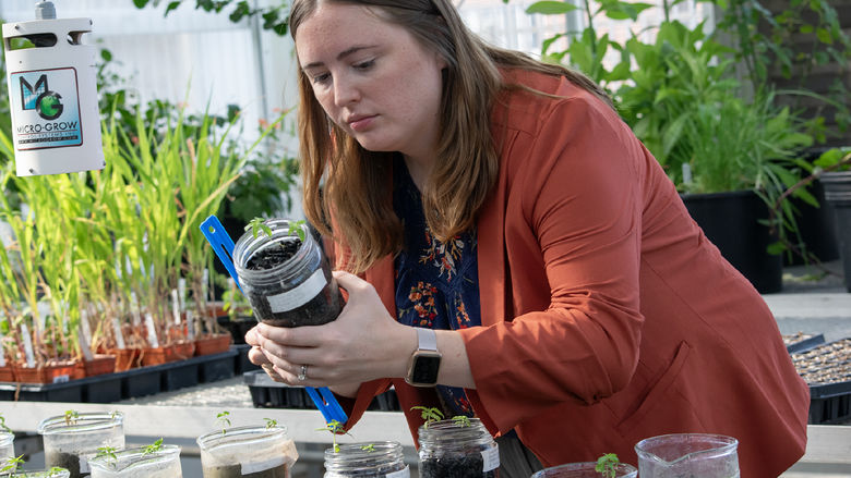 A woman holds a jar of soil and a ruler, taking a measurement, with plants behind her
