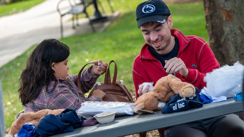 Two students sit at a table outdoors, the materials to stuff a plush animal in front of them