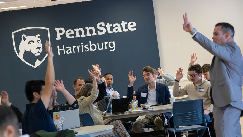 People sitting at tables raise their hands, the Penn State Harrisburg logo on the wall behind them
