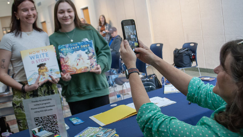 Two students pose holding children's books, while a woman takes a photo with her phone