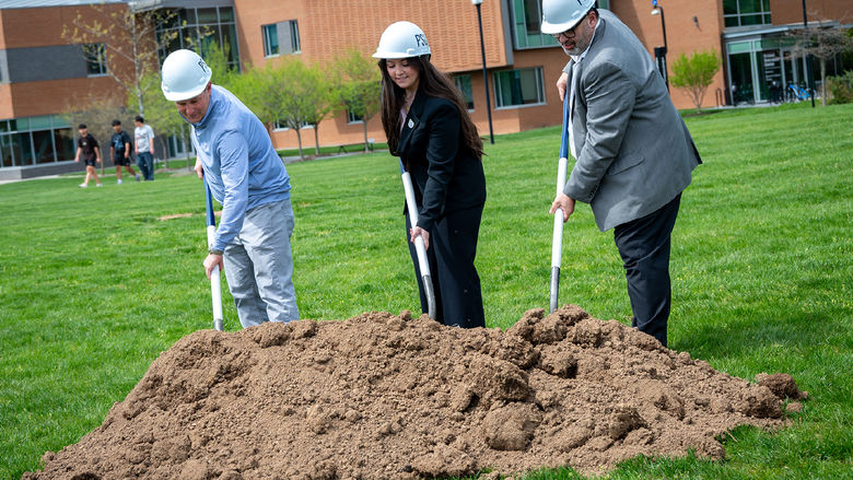 Three people wearing hard hats push shovels into a pile of dirt