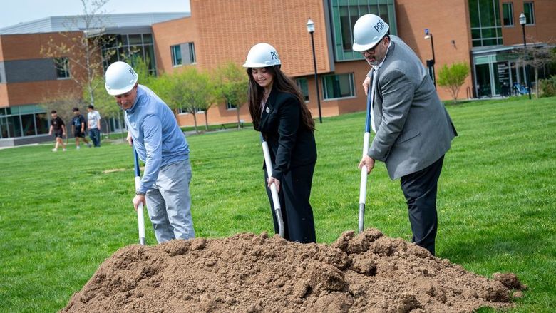 Three people wearing hard hats push shovels into a pile of dirt