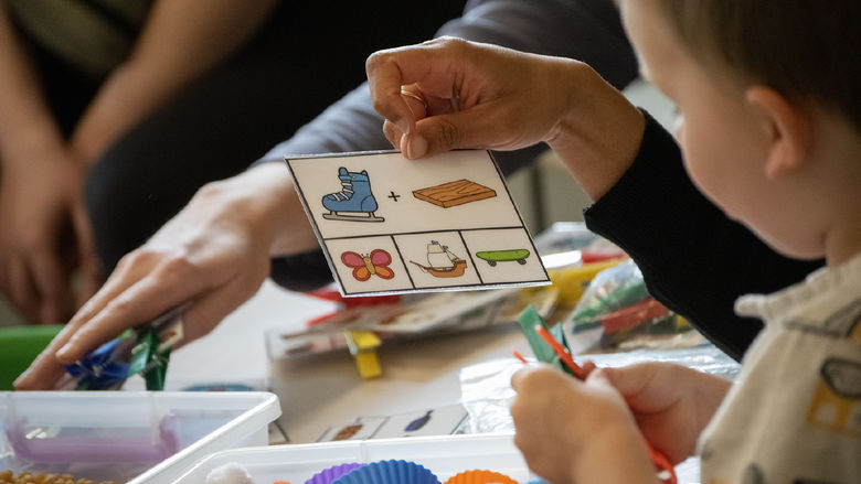 A child looks at symbols on a card, with a sensory bin of cotton balsl in front of him