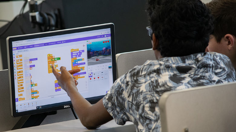 A student points to a computer screen showing a colorful coding activity