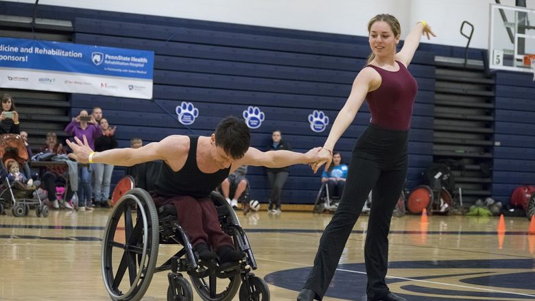 A man and a woman in leotards raise the arms on a gymnasium floor. The man in a wheelchair, bows at the waist. Behind them several people snap photos on cell phones or applaud.