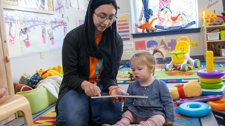 A woman reads a book to a young child in a day care center, surrounded by colorful toys