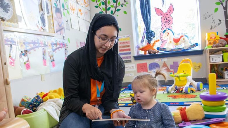 A woman reads a book to a young child in a day care center, surrounded by colorful toys