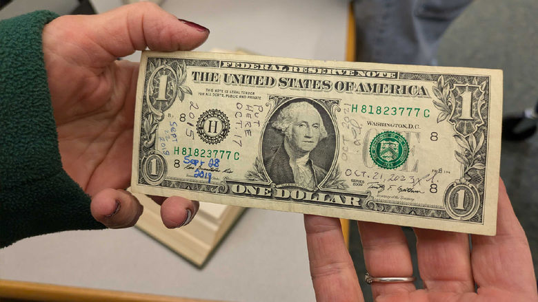 Closeup of hands holding a dollar bill. Dates are written in pen on the dollar bill
