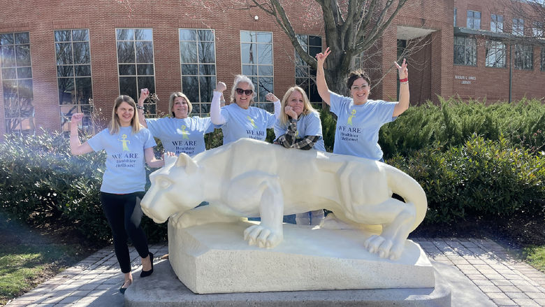A group of women in matching t-shirts strike muscle poses, standing behind the Lion statue at Penn State Harrisburg