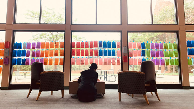 A student sits on the floor at a table in front of colorful bags taped to a large window