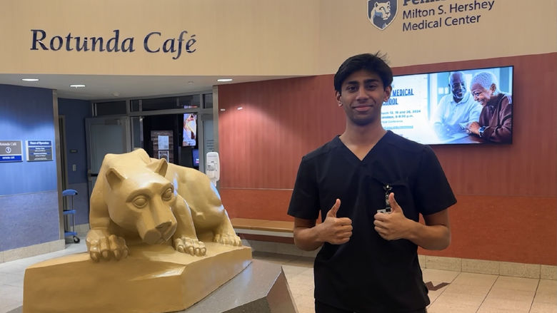 Faaiq Rizwan, wearing scrubs and giving two thumbs up, stands near a Nittany Lion statue at Penn State Health Milton St. Hershey Medical Center