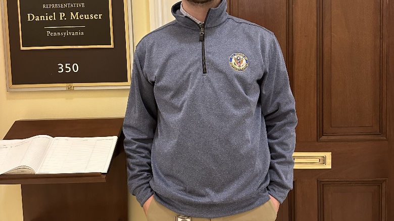 A student stands in front of an office with a sign that reads "Representative Daniel P. Meuser" 