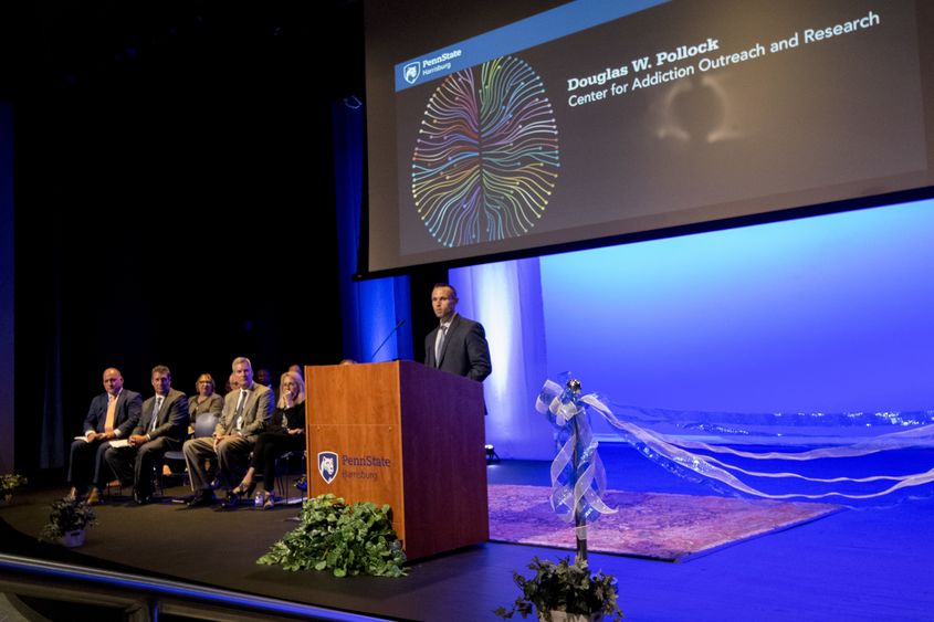 Dr. Weston Kensinger at podium, panel of speakers on his right