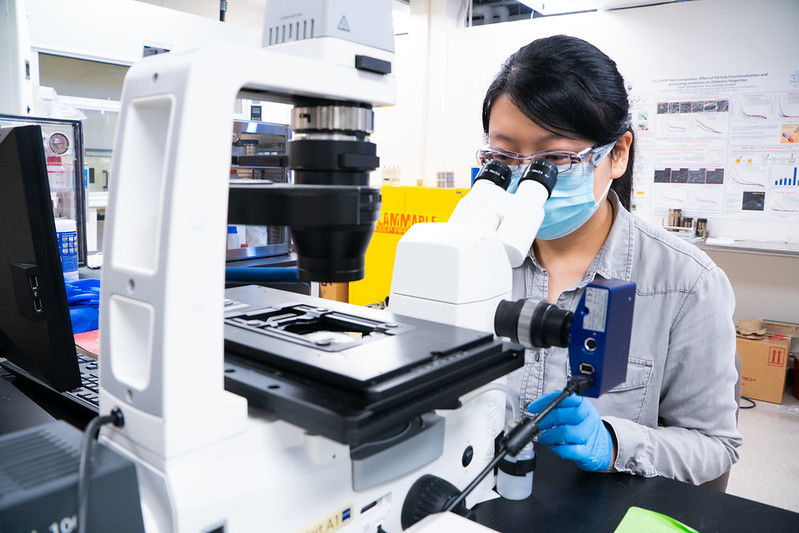 A graduate student wearing a mask looks through a microscope.