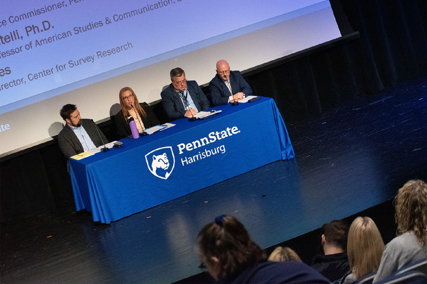 A photo of four people sitting behind a table during a presentation on a stage