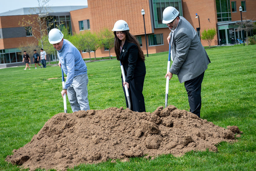 Three people wearing hard hats push shovels into a pile of dirt