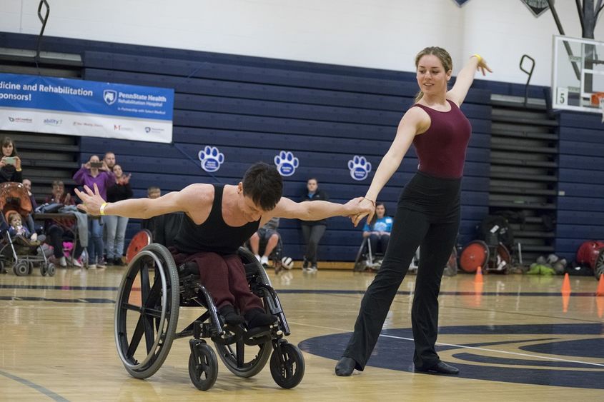 A man and a woman in leotards raise the arms on a gymnasium floor. The man in a wheelchair, bows at the waist. Behind them several people snap photos on cell phones or applaud.