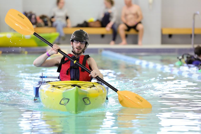 A man in an orange vest and black helmet is seated in a yellow kayak in a large swimming pool, paddling directly toward the camera.