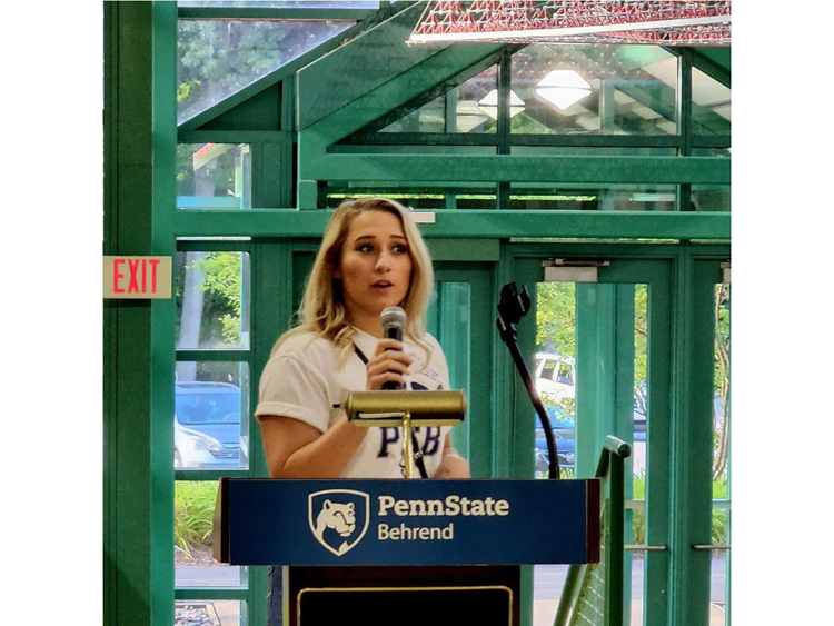 A person stands at a podium giving a speech, the podium has a banner that reads Penn State Behrend