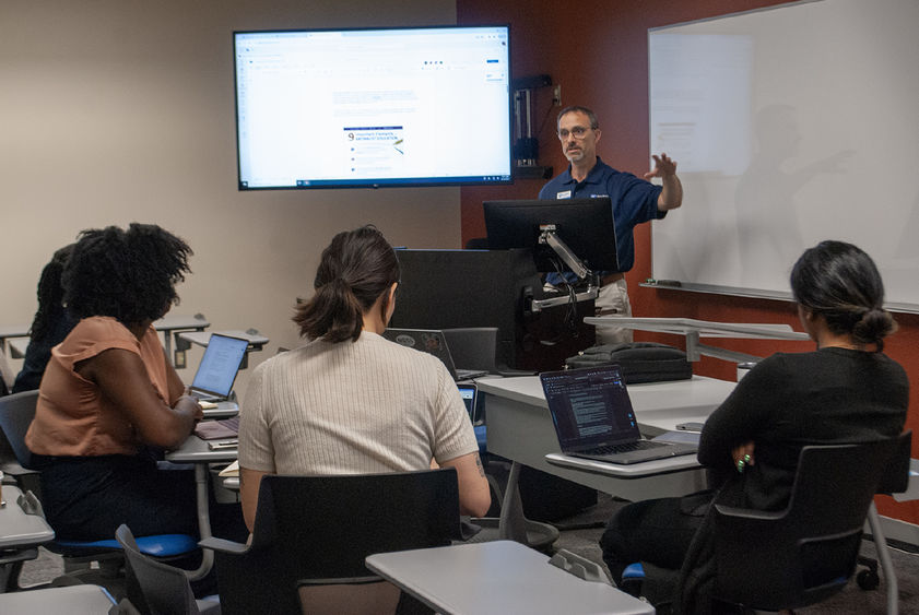 Four people sit in a semicircle in a classroom listening to speaker and watching a screen